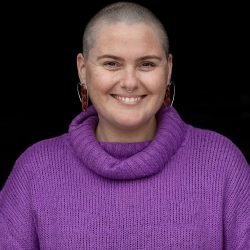 Photo of Amy smiling at the camera wearing a purple jumper and silver hoop earrings.