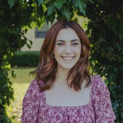 Photo of Kate, she is in a garden with lawn and trees behind her and is wearing a floral top. She is smiling at the camera.