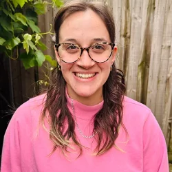 Photo of Katie, smiling at the camera and wearing a pink jumper and glasses